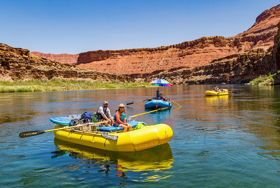 Description: yellow and blue inflatable rafts floating on a stretch of calm river water, with desert cliffs on both sides of the river. NPS/Neal Herbert