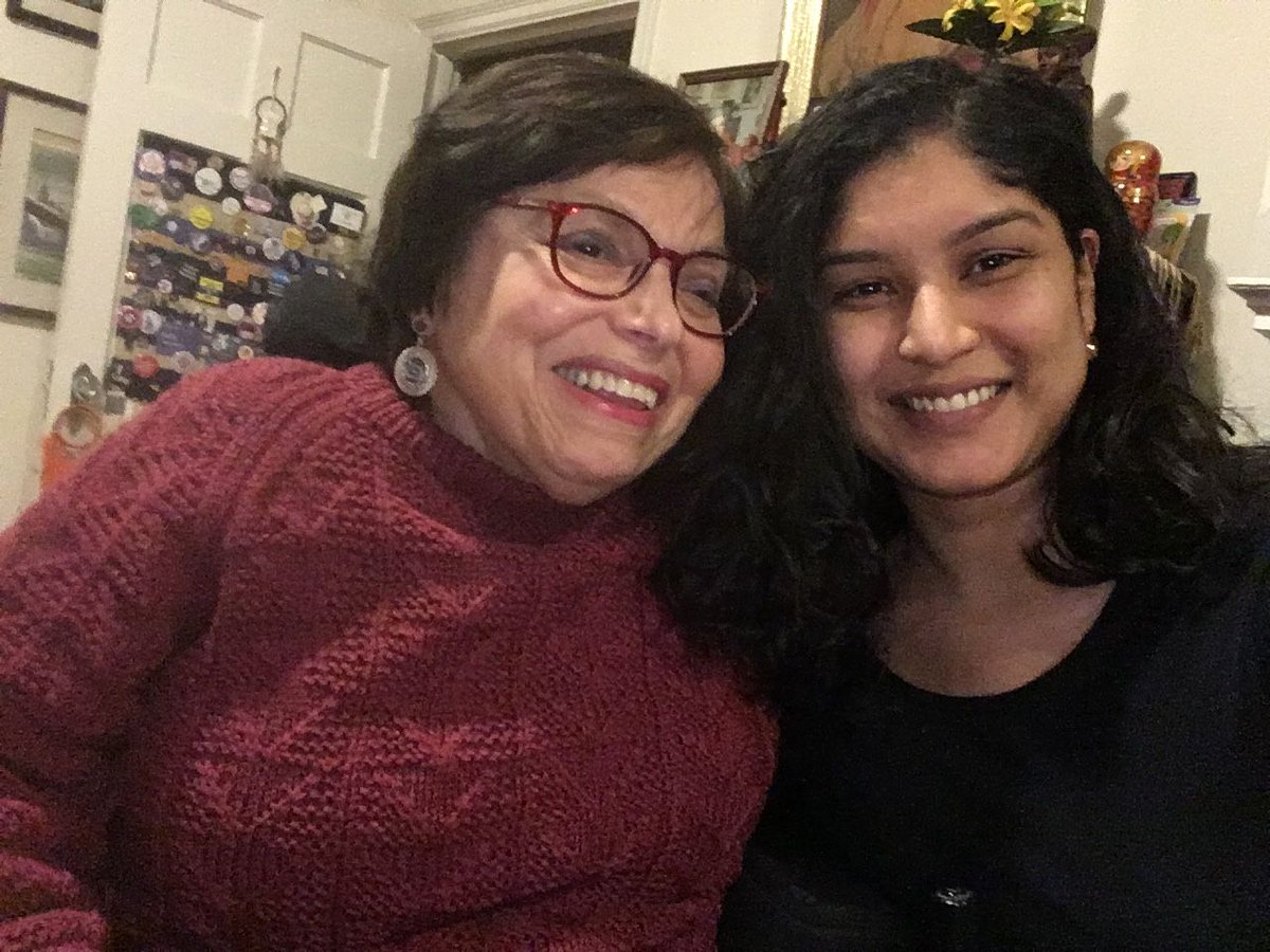Two women smiling in selfie. Woman on the left is wearing red chunky knit sweater and red glasses. She has short brown hair and circular pendent earrings. Woman on the right is wearing a black top and has large dark brown curly hair. 
