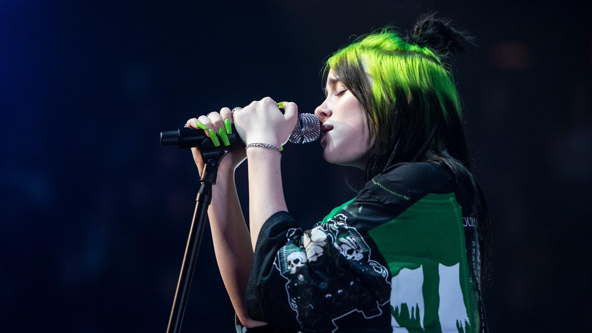 A young woman with neon green and black hair sings into a microphone.