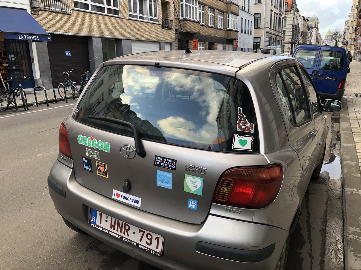 rear window of a parked gray Yaris in Brussels city center, with clouds reflected and an array of bumper stickers - including Oregon (+heart); 100% Northwest; an outline of Oregon state with a green heart inside; “a little veganism never hurt anyone”; I {heart} Europe; among others