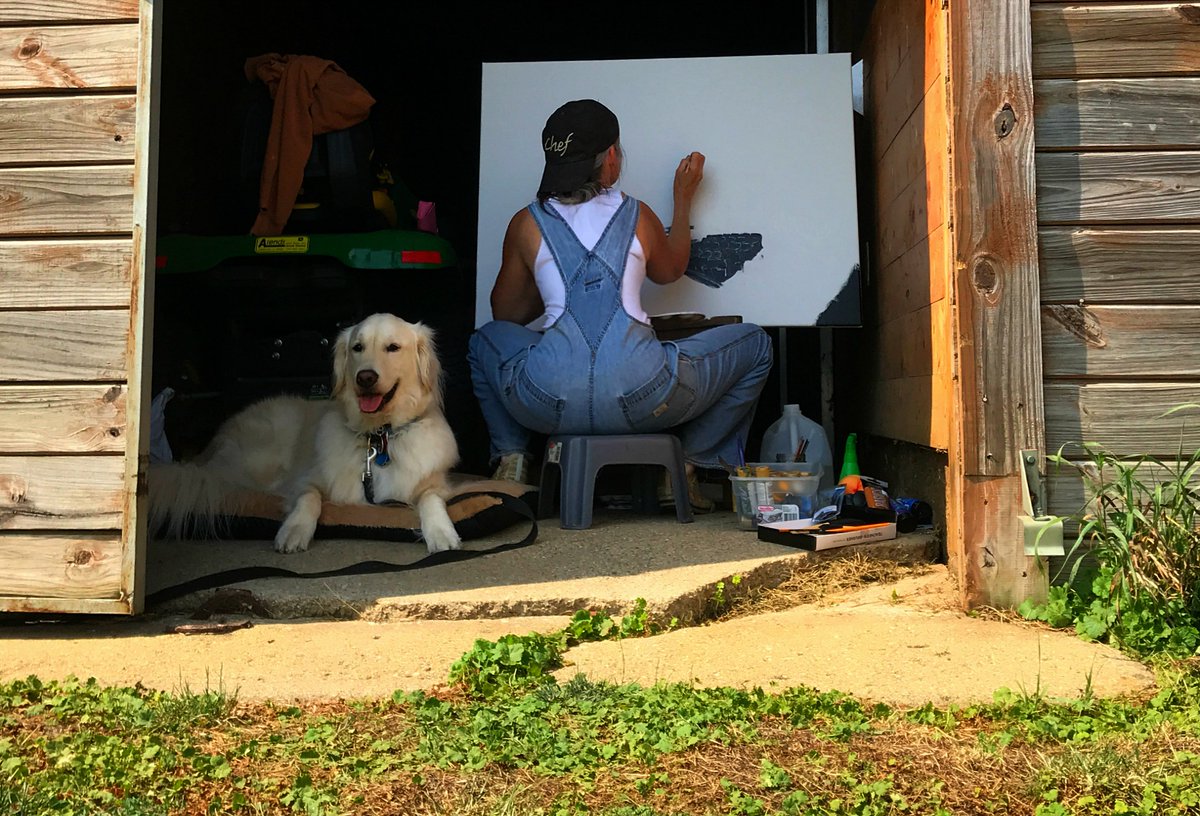 White fluffy Golden retriever dog is lying next to a person wearing blue overalls, a white tank top, and a black ball cap backwards that says "Chef". The person w/ their back to the camera and are sitting in front of a large paint canvas and they are painting. The two are in the breezeway of a barn and their is a green John Deere lawn tractor next to them. The dog looks like he is smiling. 