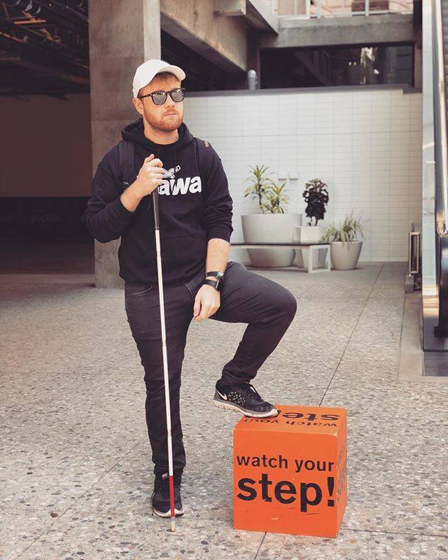 James wearing sunglasses looks neutrally confident stands with his White Cane in an outdoor shopping mall. He puts one foot on a cube block that reads "watch your step!".