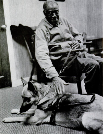 The black and white photograph shows an indoor scene with a balding man, smiling, seated in a chair, holding on to the leash of a beautiful German Shepherd guide dog with a harness. The man looks down at the dog, and the photo is evidence of the fondness between man and dog.