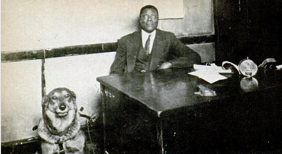 The black and white photograph shows a man in a dark suit seated at a desk, with a German Shepherd seated by the table, with his her ears pinned back, suggesting the animal is happy.