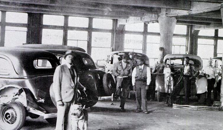 The black and white photo shows an indoor scene inside what looks like a mechanics shop in the mid-twentieth century. Men stand around the warehouse, and on the left of the image stands Herbert Douglas in a hat and suit, his hand on the harness of his guide dog.
