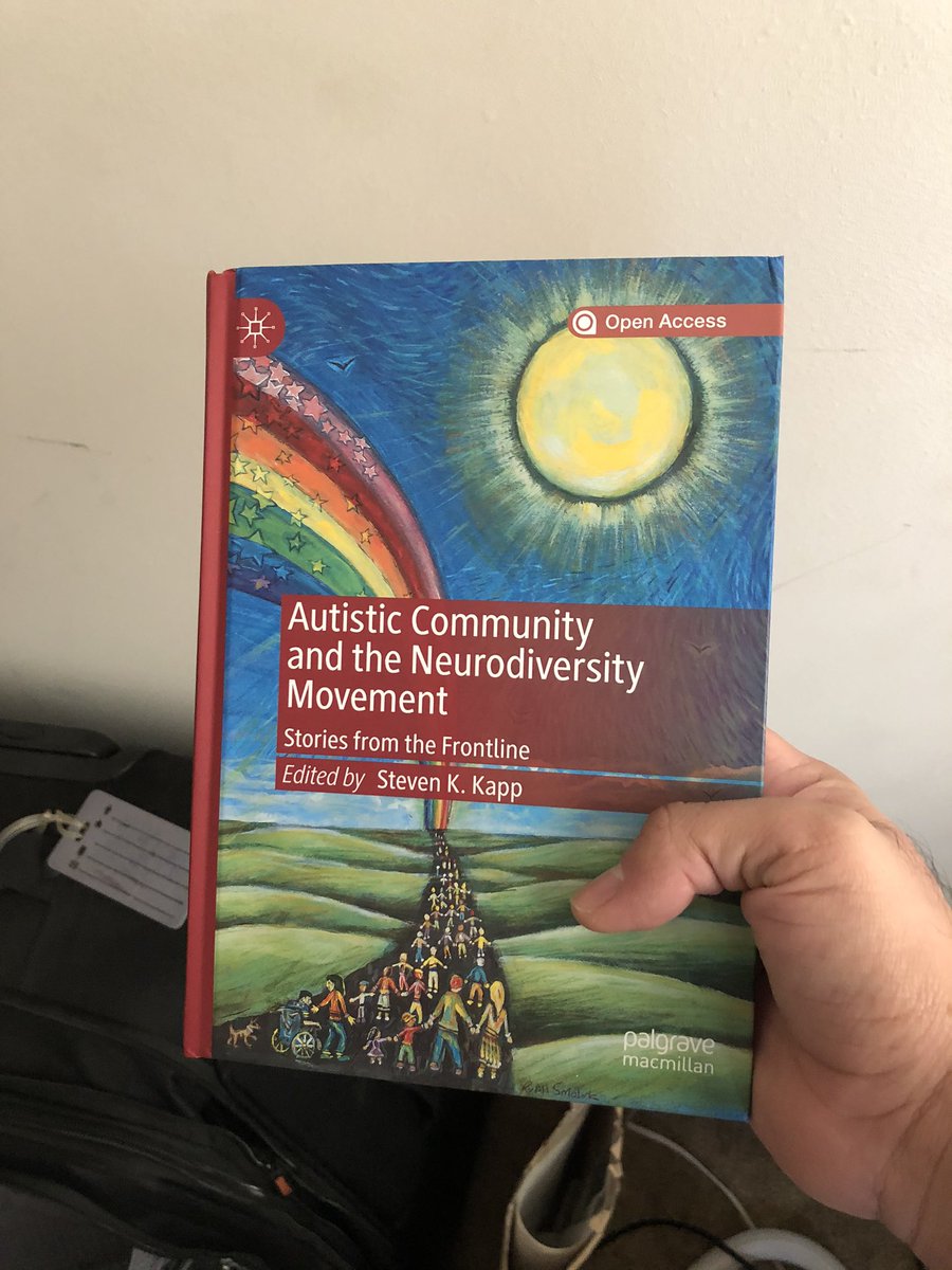 A hand holding a book with a sun and rainbow that had the title “Autistic Community and the Neurodiversity Movement: Stories from the Frontline” exited by Steven K. Kapp”