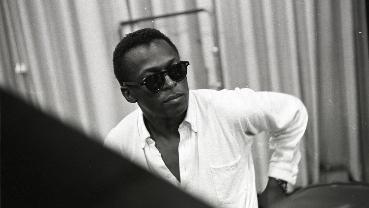 A man wearing dark sunglasses and a white button-down shirt, sits.