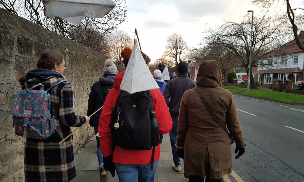 The backs of people walking with lanterns. Nearest to the camera are 3 people. Left person with a black and white coat and a floral backpack. Middle person with red coat and black back pack with a pyramid shaped lantern over his shoulder. Right a lady wearing a brown coat with the hood up.