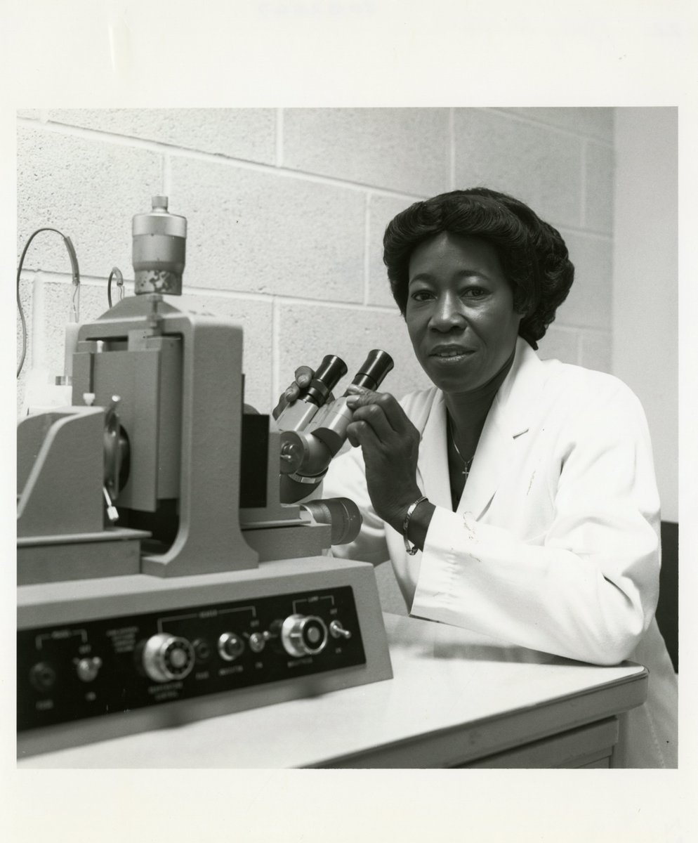 Black and white photo of an African-American woman sitting in front of a microscope. She is wearing a white lab coat.