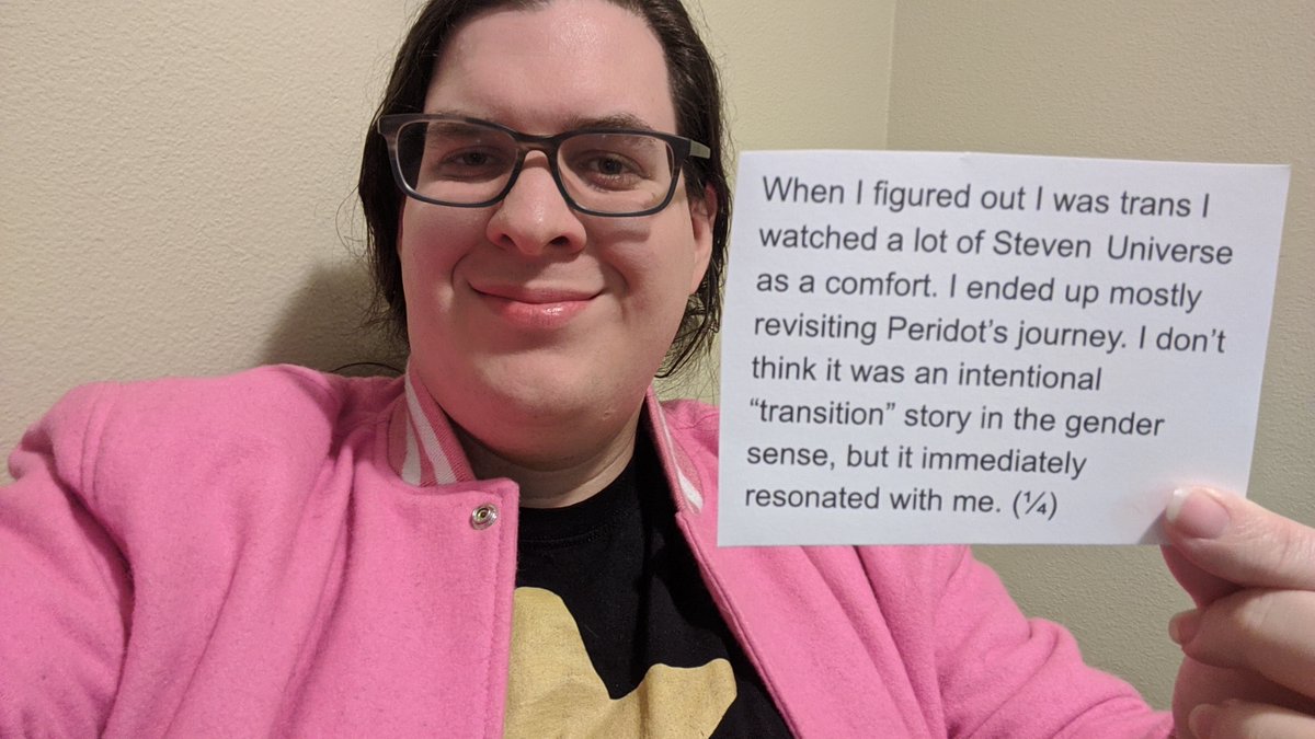 A picture of myself, holding a card saying,
"When I figured out I was trans I watched a lot of Steven Universe as a comfort. I ended up mostly revisiting Peridot’s journey. I don’t think it was an intentional “transition” story in the gender sense, but it immediately resonated with me. (¼)"
I am wearing a pink jacket and black shirt with a star (like Steven Universe in SU Future).