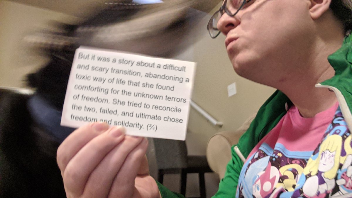 A picture of myself, holding a card saying,
"But it was a story about a difficult and scary transition, abandoning a toxic way of life that she found comforting for the unknown terrors of freedom. She tried to reconcile the two, failed, and ultimate chose freedom and solidarity. (²⁄₄)"
I am wearing a pink shirt and green jacket and kissing a very blurry dog in my face