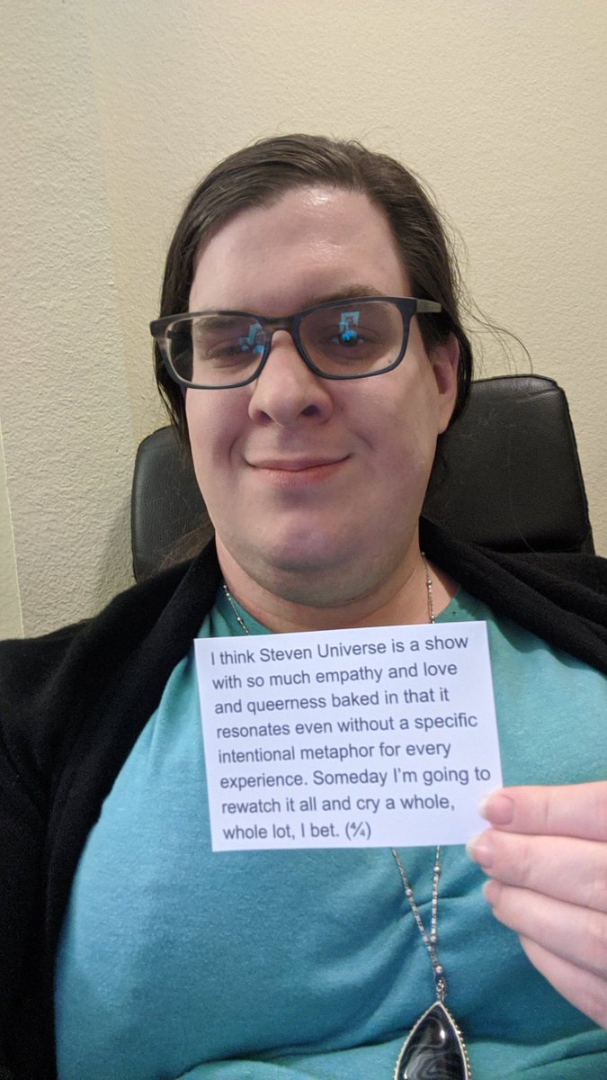 A picture of myself, holding a card saying,
"I think Steven Universe is a show with so much empathy and love and queerness baked in that it resonates even without a specific intentional metaphor for every experience. Someday I’m going to rewatch it all and cry a whole, whole lot, I bet. (⁴⁄₄)"
I'm weaing a teal shirt, teal glasses, blue cardigan, and a large teardrop-shaped pendant.