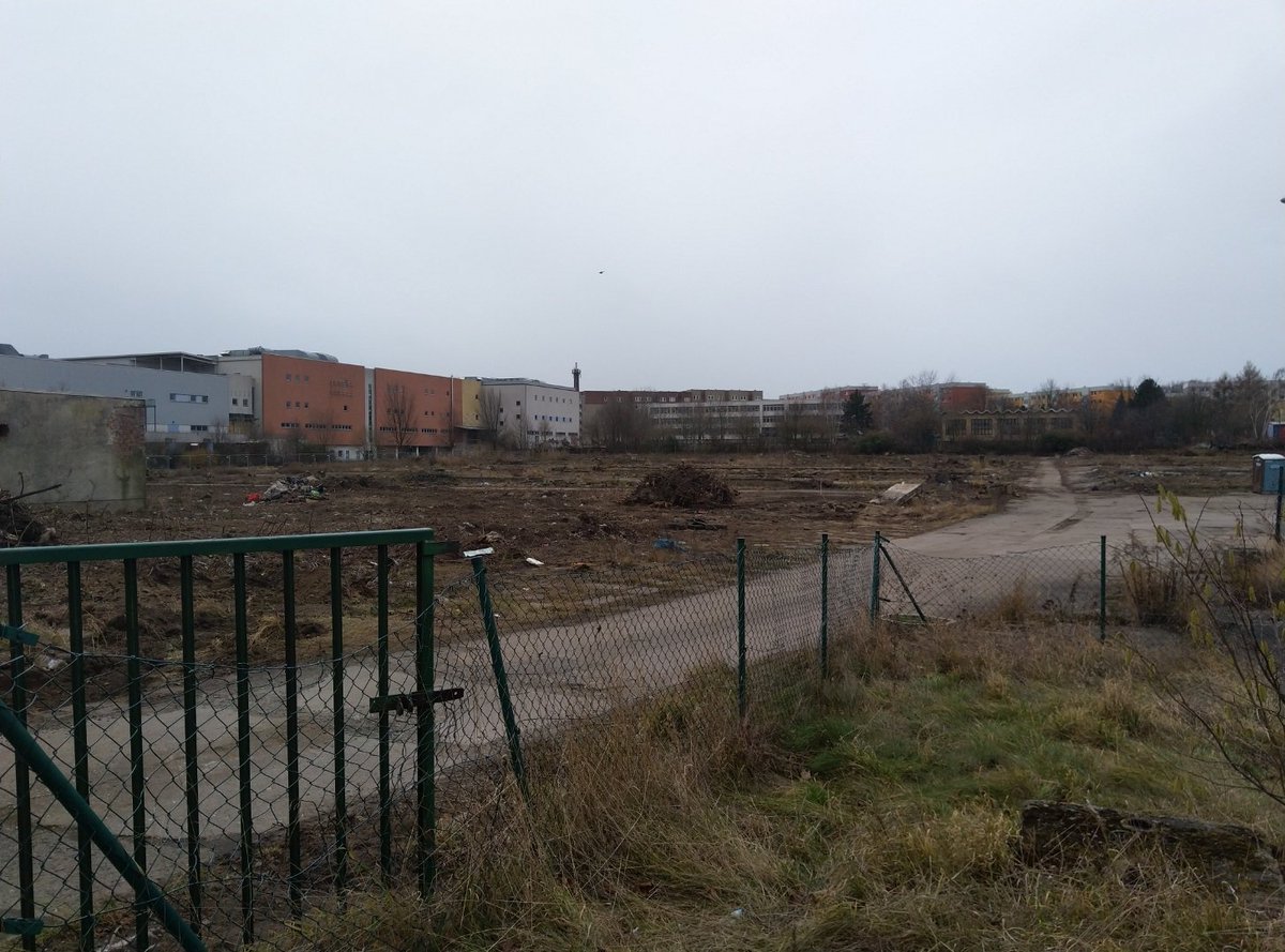 Eine karge Fläche, Boden liegt frei, alte Grundmauern und viel Müll und ein paar Getrüpreste, ein kaputter Maschendrahtzaun. Im Hintergrund Mehrfamilienhäuser.