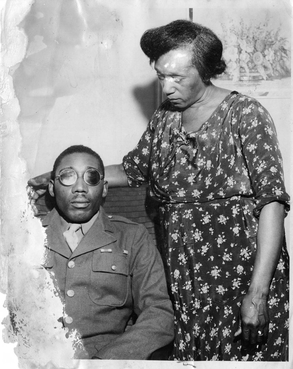 The black and white photo shows Isaac Woodard in uniform seated with dark round glasses with his mother standing next to him, in a darkish dress with floral print.