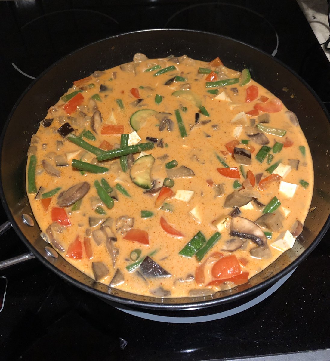 A wok on a stove filled with a thin reddish sauce and a lot of vegetables (mushrooms, zucchini, green beans, red peppers) and tofu
