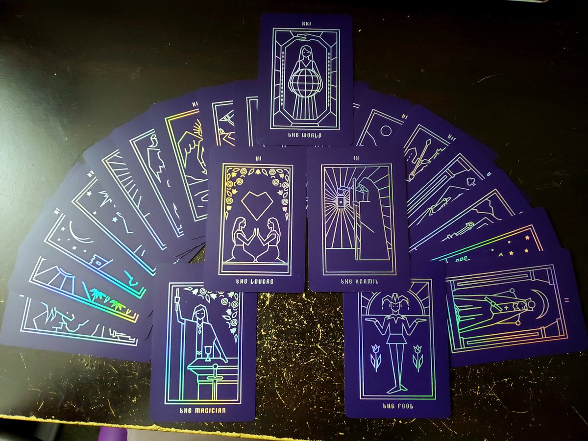A semi-circular spread of the tarot cards with The World, The Lovers, The Hermit, The Magician, and The Fool laid overtop in a triangular pattern. The text and line art on all the cards is the same holographic material used in the box.