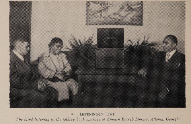 The black and white photograph shows three people, two men and one woman seated around a desk with a machine on it. Two potted plants sit next to the talking book machine and a picture hangs on the wall behind the desk.