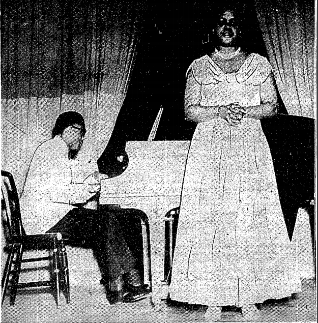 The image shows a grainy black and white photograph of a woman in a full length gown with dark hair and a necklace around her neck, standing on stage, singing with her hands clasped. behind her, a seated man with glasses plays the piano.