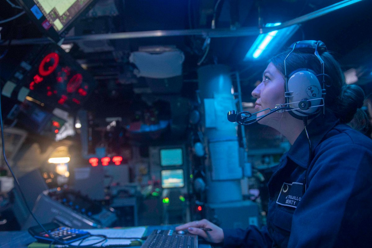 A Sailor looks up at a monitor as she works on a computer system on a ship.