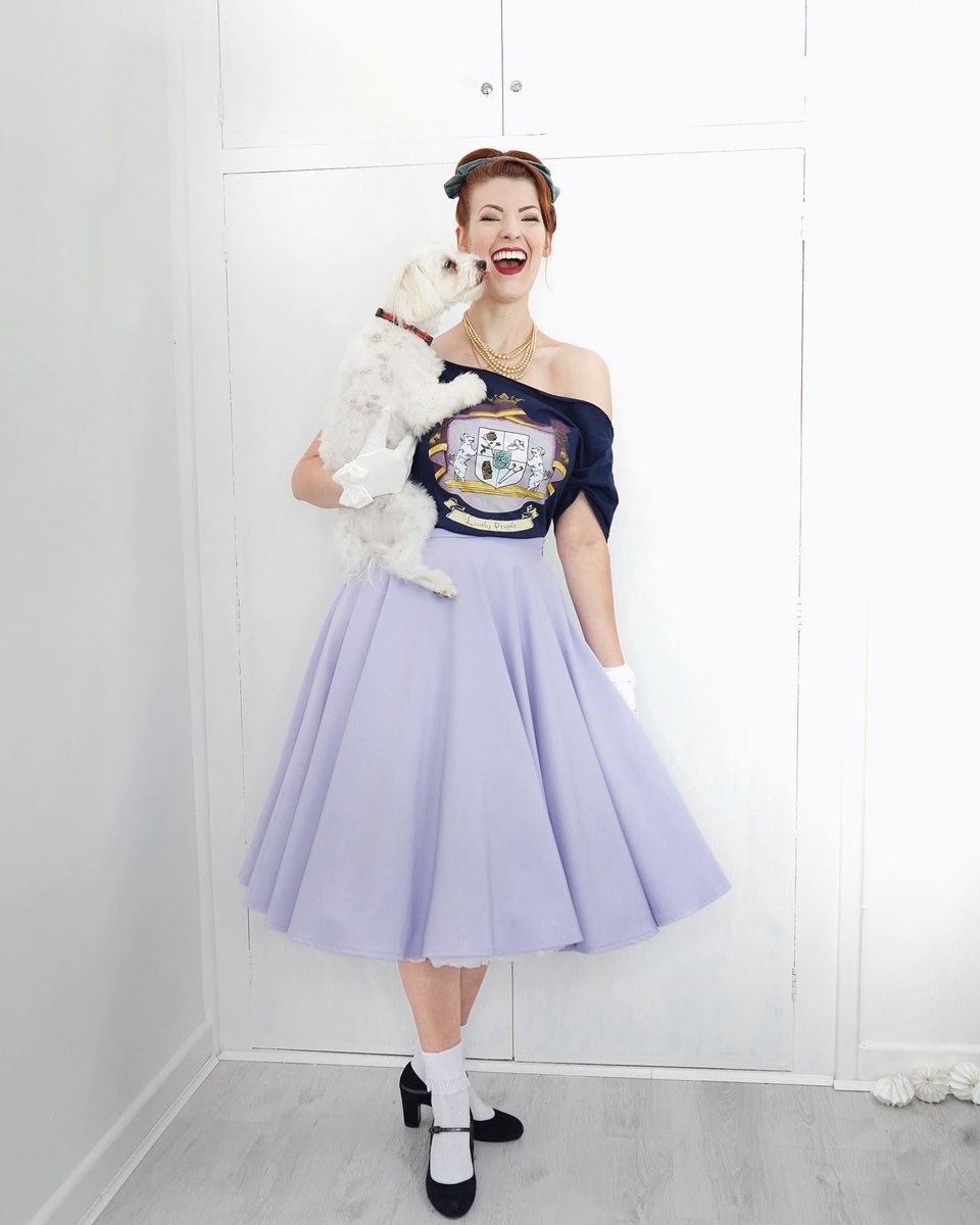 Jessica holding Tilly, who is licking the side of Jessie's face. Jessica is in a navy lovely people shirt resting on her shoulders paired with a lilac skirt and black heels. 