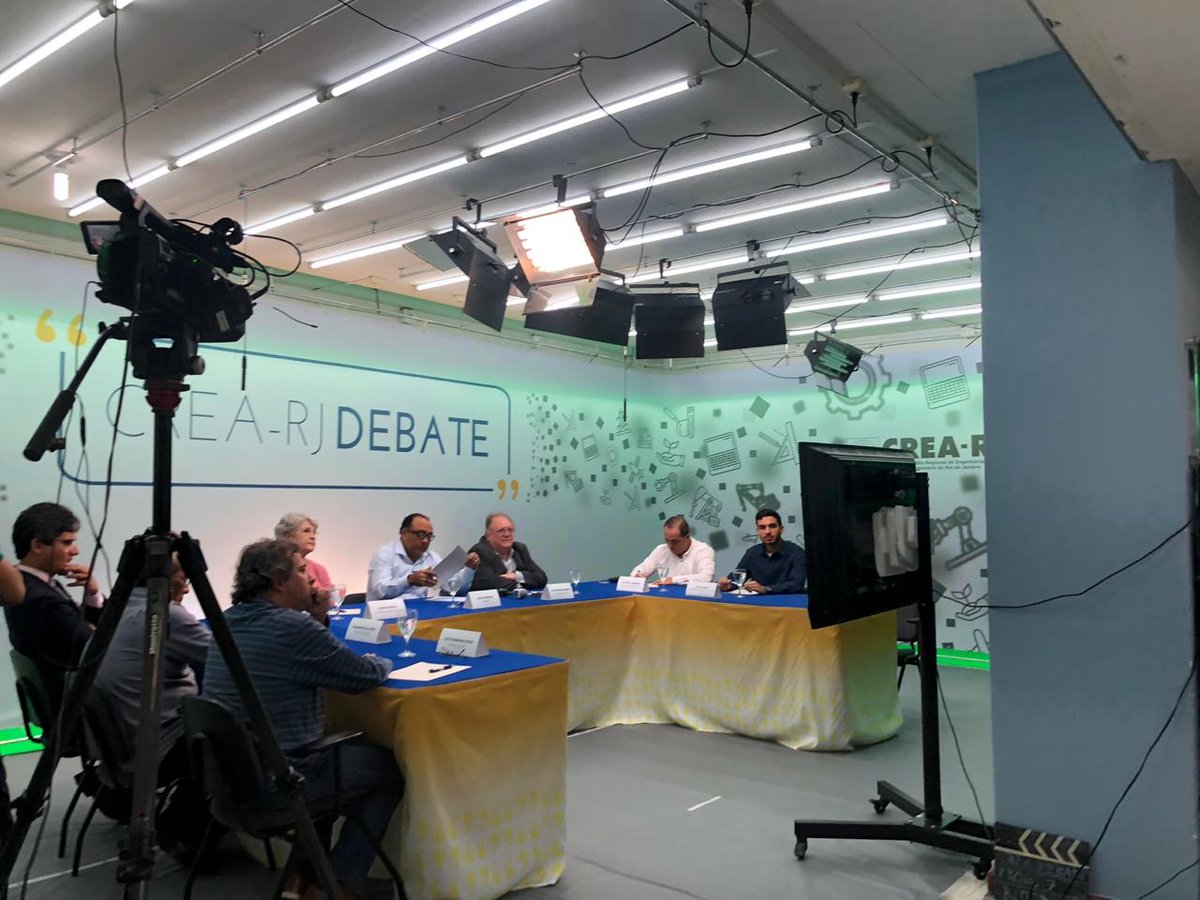 Team members sitting at tables with lights and cameras facing them, presenting their work.