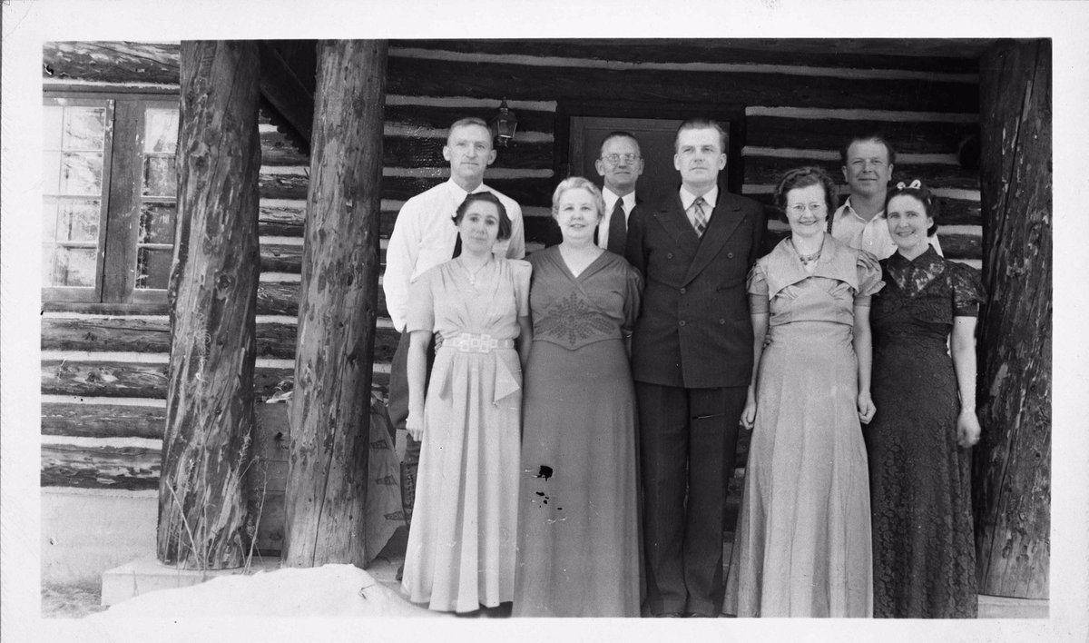 Eight people stand in front of a log cabin. There is snow on the ground. Men are wearing suits and ties; women are wearing short-sleeved ankle-length dresses. Denali National Park and Preserve Museum Collection, DENA 22659.