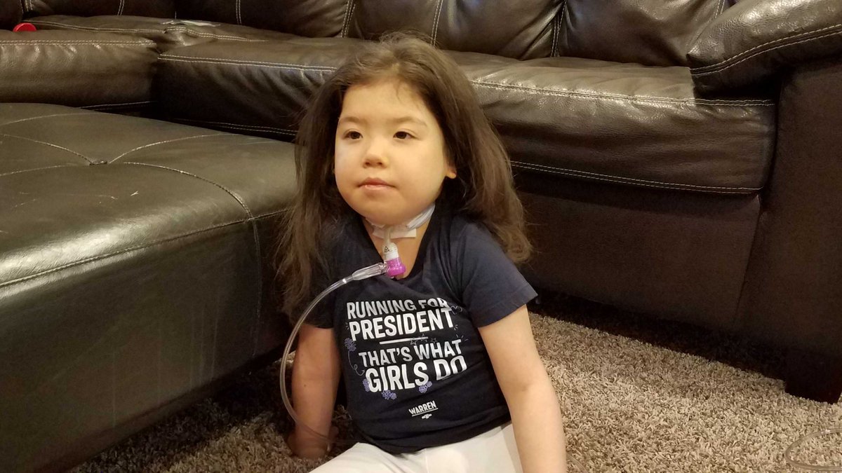 photo of Xiomara, young girl with trach (connected to oxygen tubing) and hair down wearing a blue Warren shirt that says “Running for president - that’s what girls do