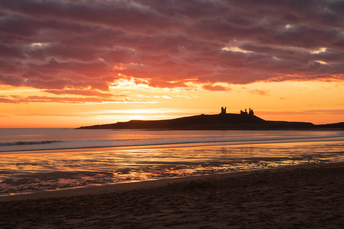 Blazing orange sunset with the silhouette of Dunstanburgh Castle in the background and a beach in the foreground.