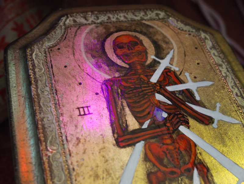 Painting of red skeleton on gold leaf background, pierced with white swords. The skull is darkly haloed. A three in roman numerals is painted to the left of the figure. The painting is varnished in gloss, and reflects fuchsia ambient lighting.