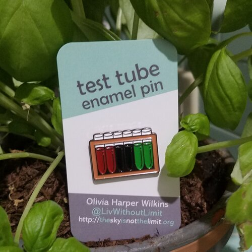 Test tube pin with two dark red, two black, and two green test tubes on a mint green, white, and grey backing card resting in front of a basil plant.