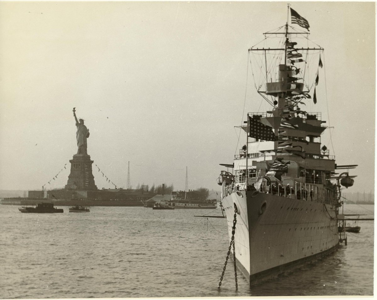 A black and white photo of a large military ship with the Statue of Liberty in the background.