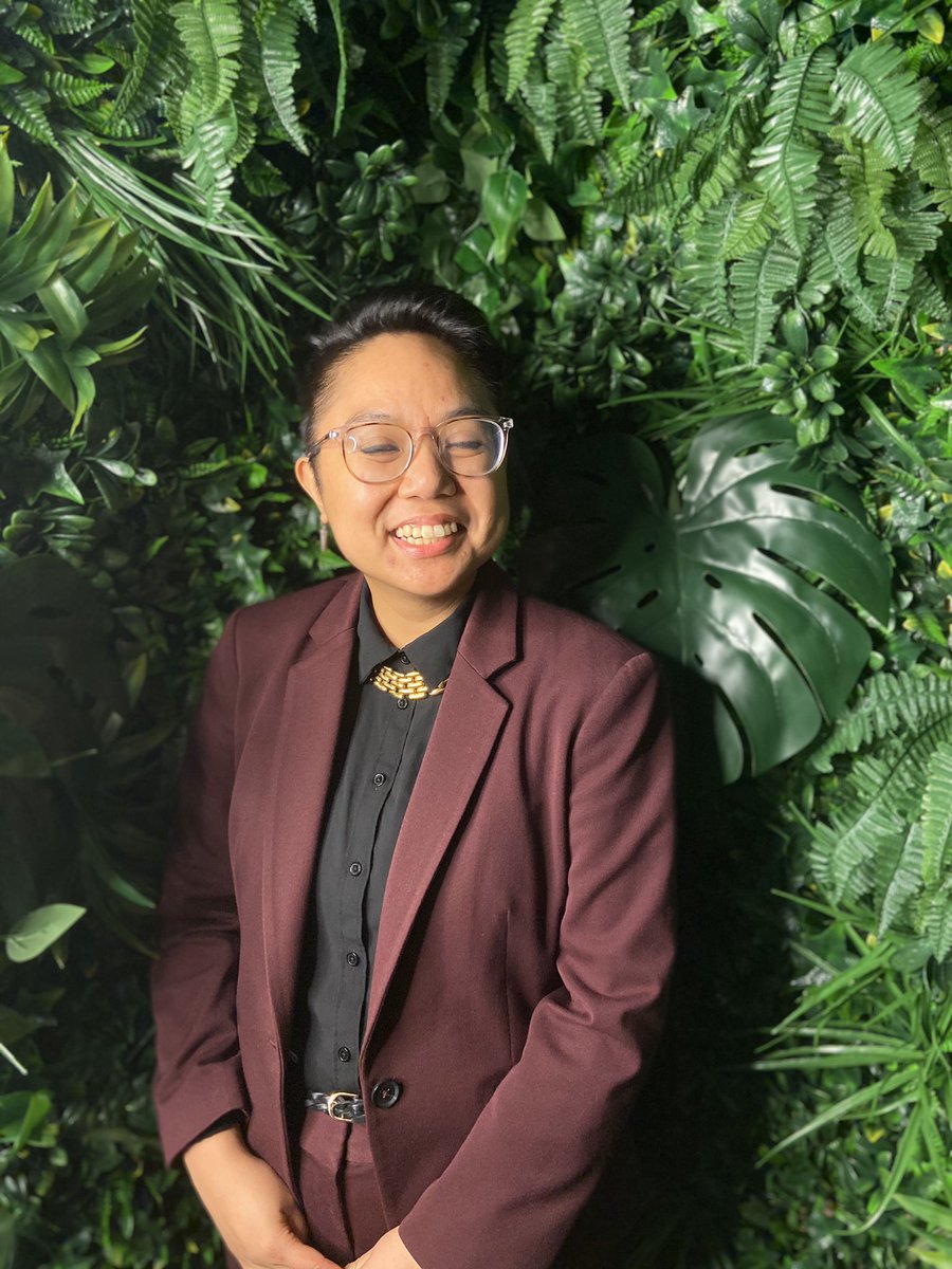 Portrait of AC in a maroon suit in front of a green plant wall. They are smiling and have their eyes closed