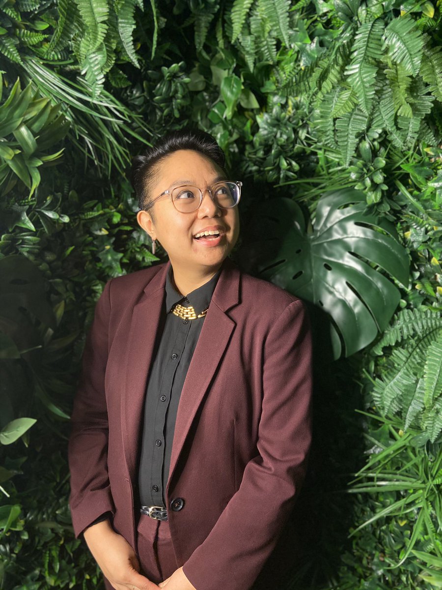 Portrait of AC in a maroon suit in front of a green plant wall. They are smiling and looking up to the right