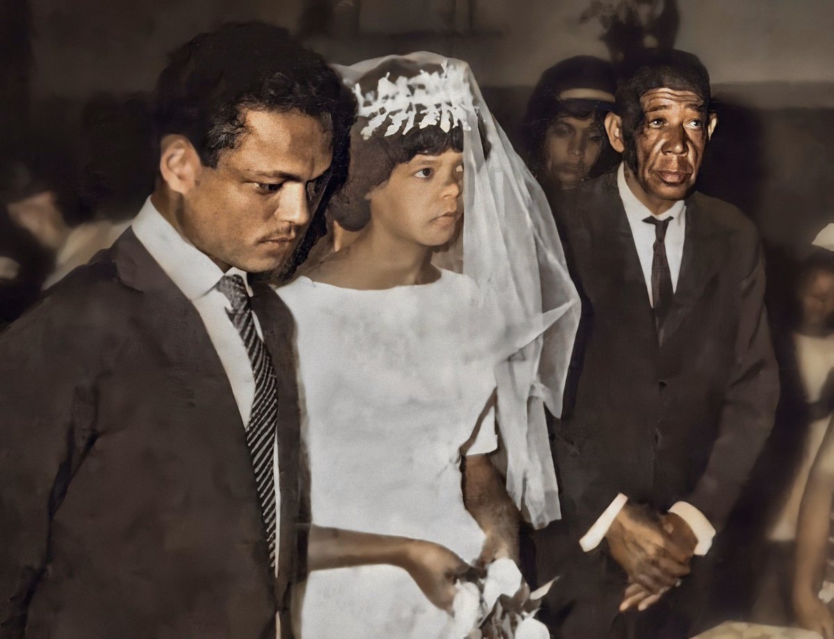 Cena de casamento, 3 pessoas em destaque olhando para o altar. Um jovem homem de terno e gravata pretos e camisa branca; uma jovem de vestido de noiva branco com véu na altura do cotovelo; um senhor já de aparência idosa de terno e gravata pretos e camisa branca. Todos são negros de pele escura, sendo o senhor idoso o de pele mais escura no grupo.