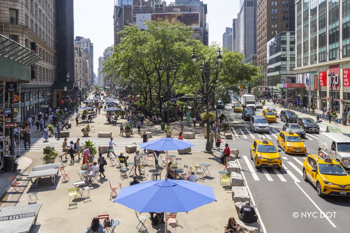 An aerial view of a pedestrian plaza on a sunny day. Cars are driving alongside the plaza while some people are sitting in chairs and some people are walking across the pedestrian plaza. 

