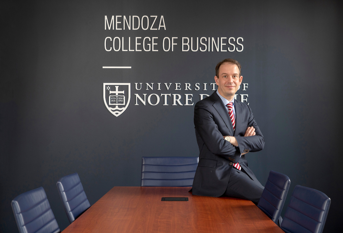 A man wearing a suit leans on a boardroom table with his arms crossed in front of a sign reading Mendoza College of Business University of Notre Dame