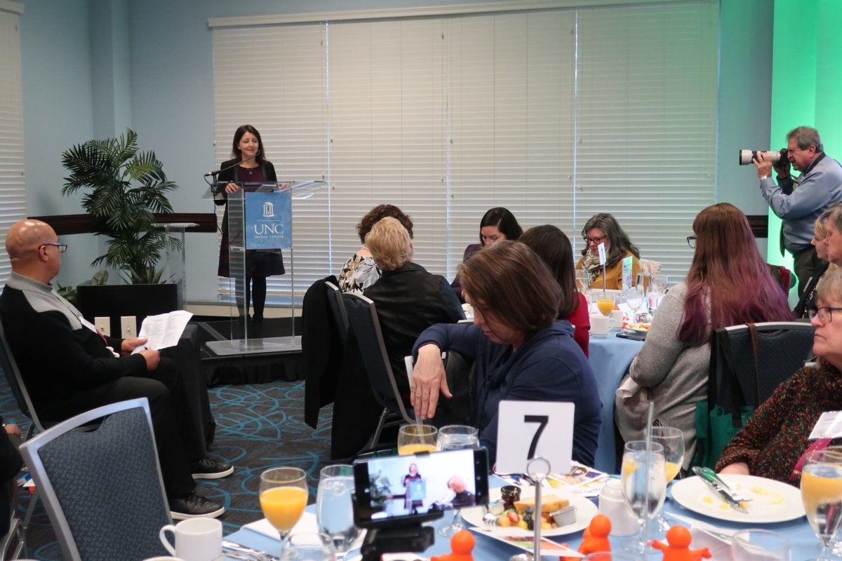 A woman at a dais speaking to audience members over breakfast