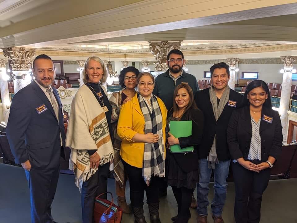 8 people stand and smile for a photograph in a legislative chamber gallery.