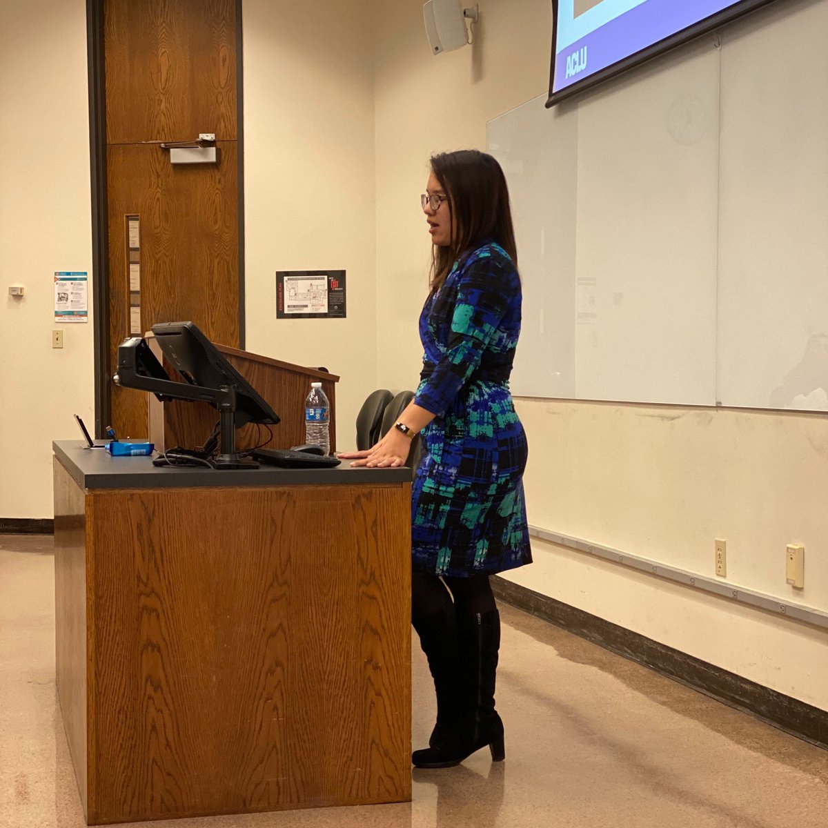 Image: ACLU of Texas attorney Anjali Salvador leans on long desk in a classroom. She addresses an audience that is not pictured and has a powerpoint presentation projected on a screen behind her.  