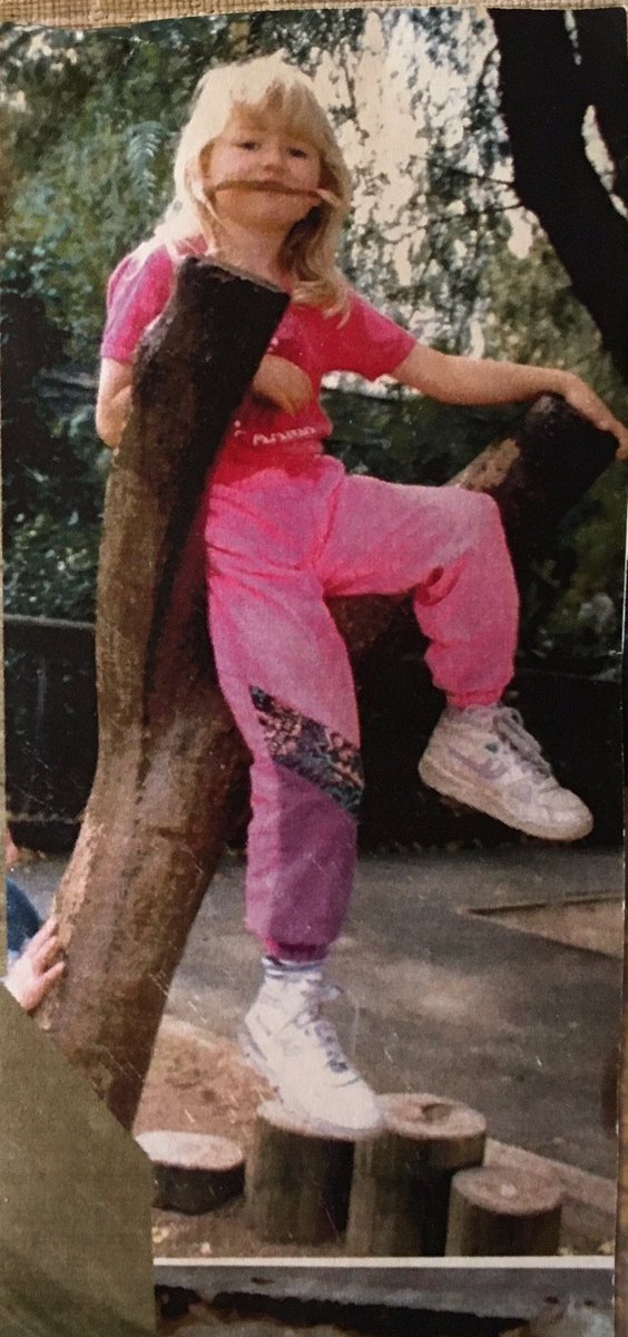 A blonde haired girl sits in the fork of a tree with a gun leaf in her mouth. She’s wearing fluoro pink pants and T-shirt and weirdly large white hi-top sneakers.