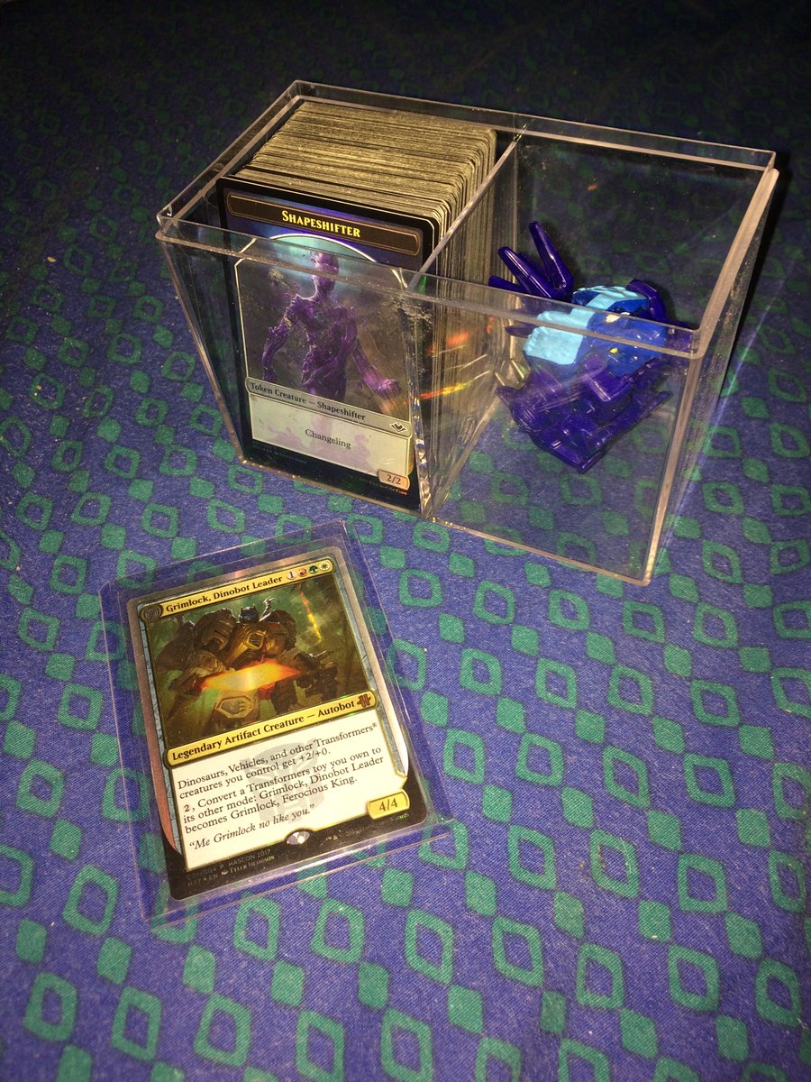 Plastic box holding one deck, a foil shapeshifter token visible, and a tiny Transformer stegosaurus. In front, in hard case sleeve, is the MTG card Grimlock, Autobot Leader