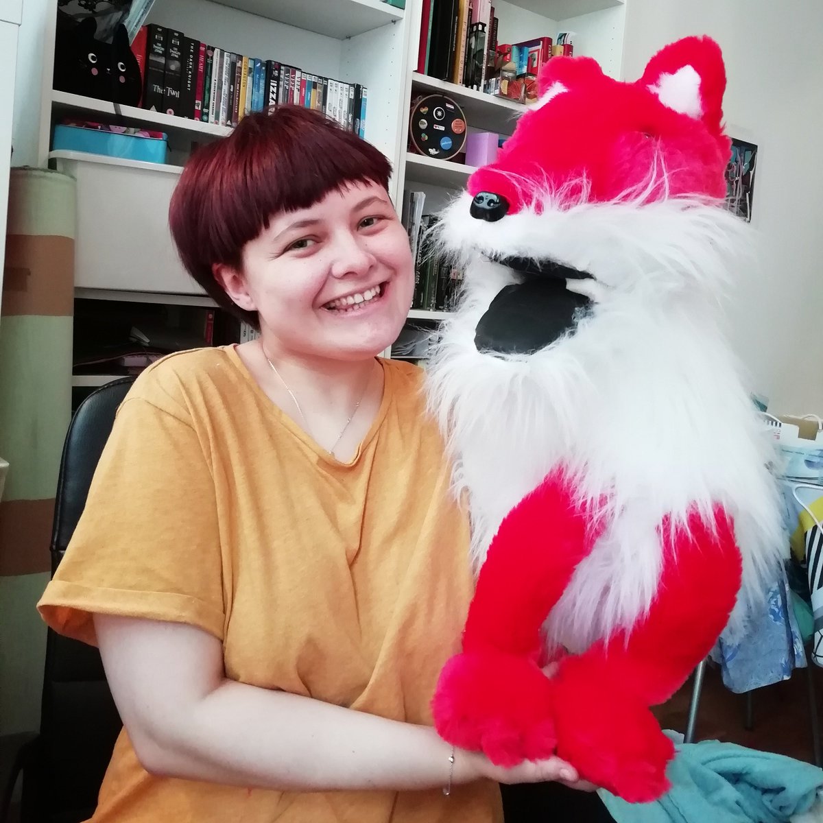 Girl with short red hair wearing a yellow top, holding a puppy fox in her left hand. The fox is red and fluffy, with white chest and mouth. They are both smiling to the camera