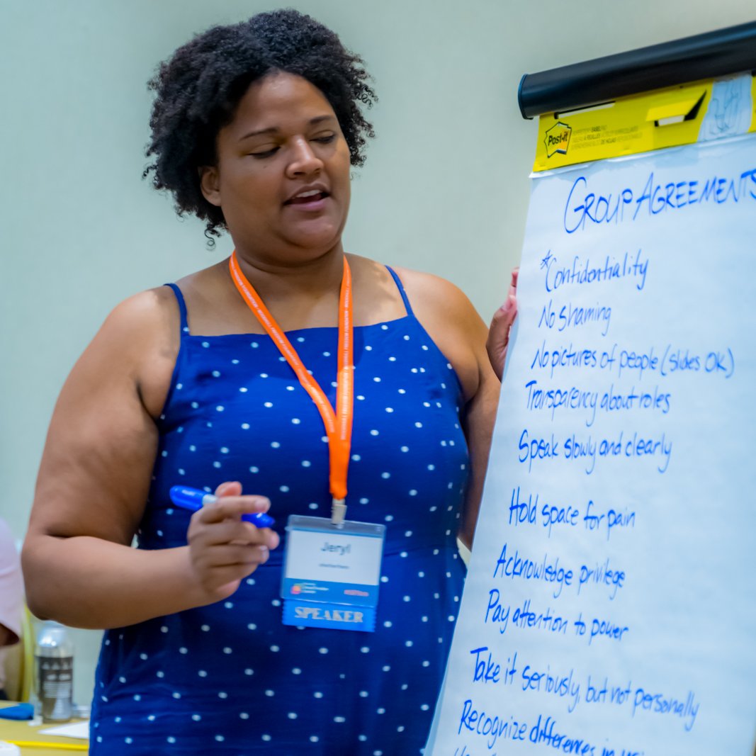  A presenter stands with a large white pad of paper with the words "Group Agreements" at the top.