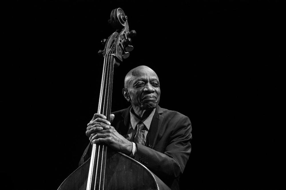 Jazz musician Reggie Workman who is an older African American man. In this photo he is dressed in a suit and holding his bass by the staff. The photo itself is in grayscale.