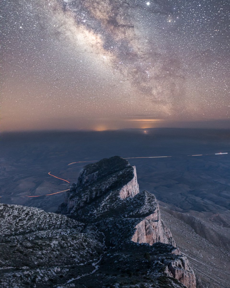 A view from a rocky outcropping down to a bare, flat desert plain far below with a bright, starry sky above.