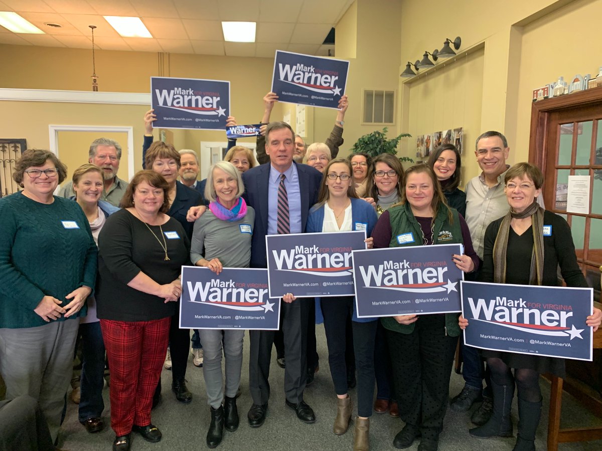 Mark Warner standing with committee chairs, vice chairs and former candidates in Warren County, VA. Several people are holding Warner campaign signs.