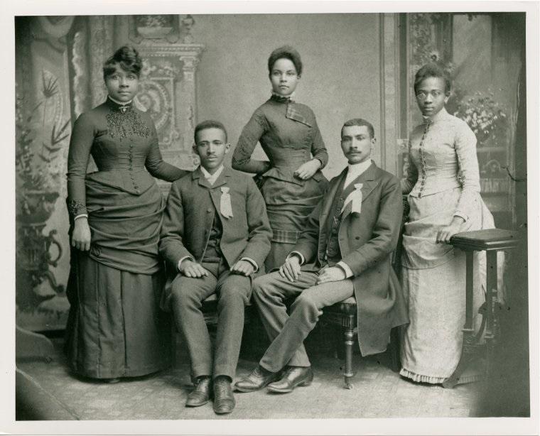 W.E.B. Du Bois posing with 4 other Black graduates of Fisk University in 1888. Three women stand around the two seated men (including Du Bois on the left).