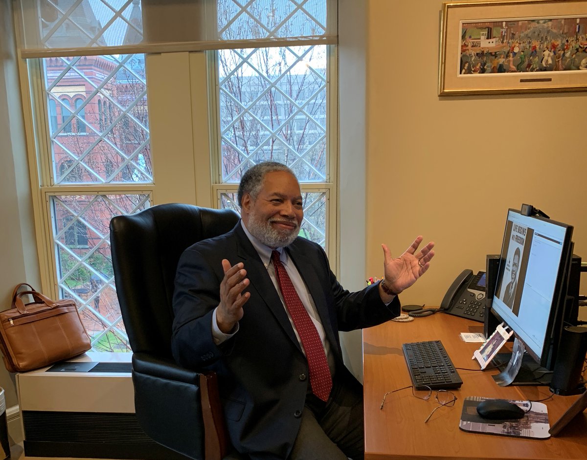 Lonnie G. Bunch III is seated at his desk. His hands are raised in a “we did it, voila” gesture and he smiles. Behind him, his satchel is placed on his windowsill. Outside the window, a view of the Smithsonian Castle Garden, Castle, and Arts and Industries Building.