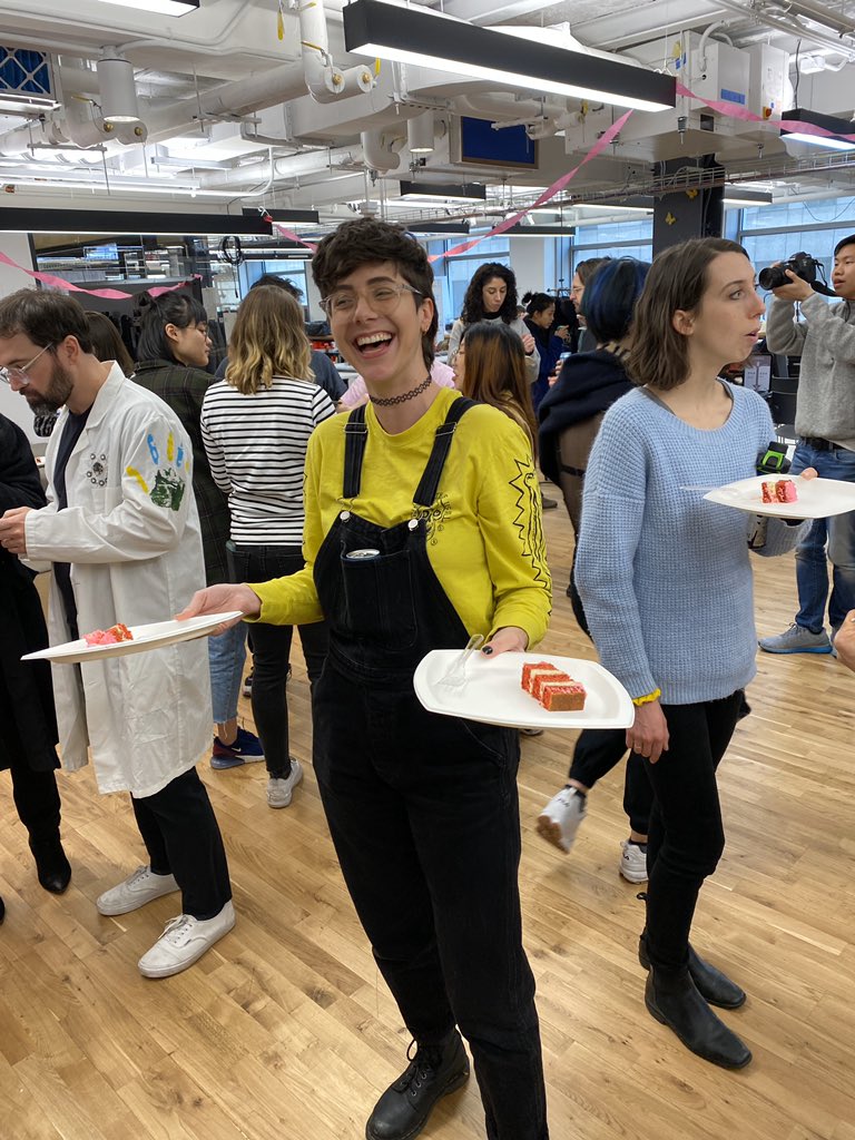 Cassie stands holding two pieces of red velvet cake with lots of itp students surrounding the background!