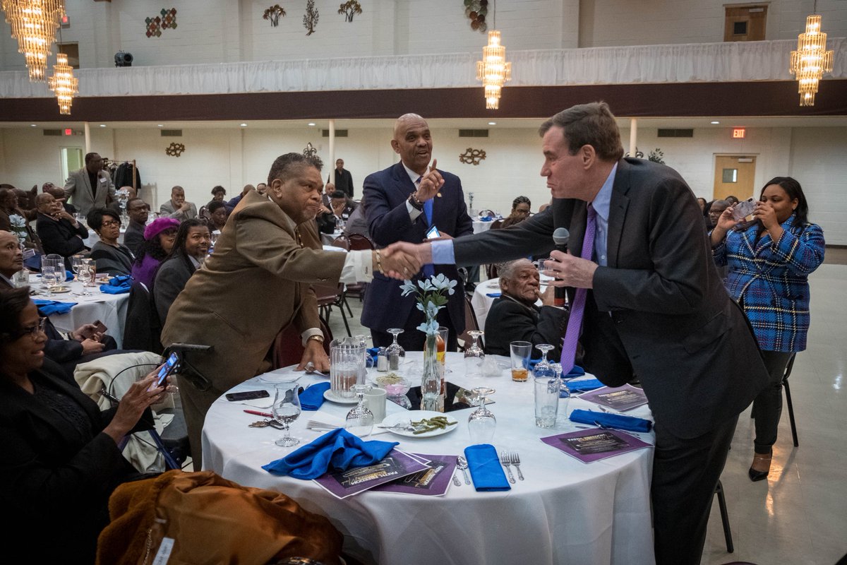 Senator Mark Warner leans over a table to shake another man's hand.